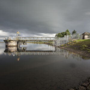 Under a heavy, dramatic overcast sky, the glassy expanse of the reservoir mirrors the precise geometry of the Victorian ironwork bridge and intake tower. Long exposure softens the surface into a mirror-like sheen, contrasting against the textured stone bank and the quiet, solitary utility house on the shore. It is an exploration of industrial heritage framed by the vast, contemplative quiet of the Scottish landscape. Photographer: Stephane Loustalot