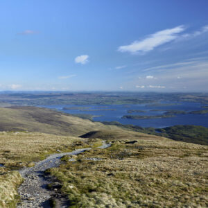 Looking out from the high ridge, a rugged stone path guides the eye downward into a vast, island-dotted expanse of water below. The composition balances the textured, weathered foreground of the uplands with the serene, luminous blue of the sky, offering a profound sense of openness and tranquility. Photographer: Stephane Loustalot