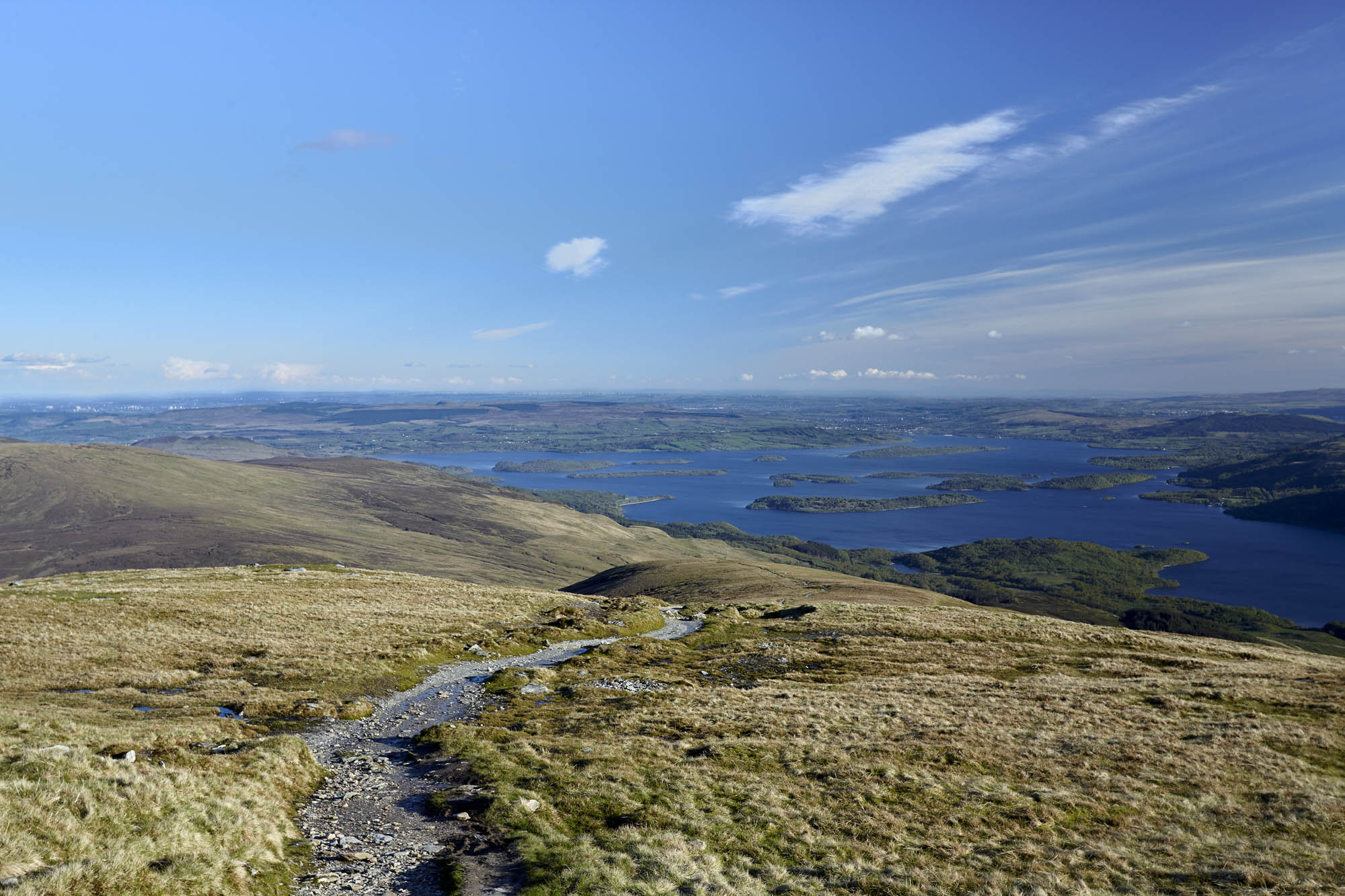 Looking out from the high ridge, a rugged stone path guides the eye downward into a vast, island-dotted expanse of water below. The composition balances the textured, weathered foreground of the uplands with the serene, luminous blue of the sky, offering a profound sense of openness and tranquility. Photographer: Stephane Loustalot